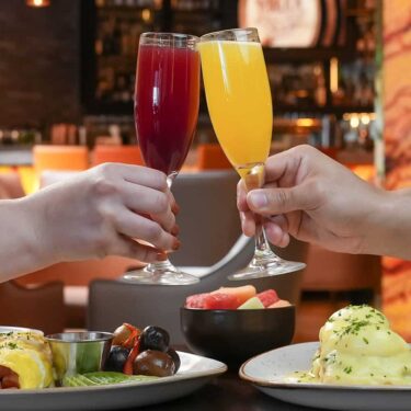 Two people toasting with glasses filled with red and orange cocktails at a brunch table with plates of eggs benedict and fresh fruit.