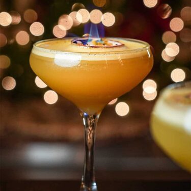 A flaming cocktail in a stemmed glass with a blurred background of golden bokeh lights at a whiskey bar. A slice of citrus garnishes the drink, enhancing the visual appeal of the fiery presentation.