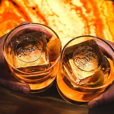 Two glasses containing amber-colored drinks with ice cubes engraved with "rocks" rest on the polished counter of a whiskey bar. The glasses are held against a warm, glowing orange background. Each glass is
