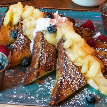 A plate of french toast topped with powdered sugar, fresh berries, a dollop of cream, and a side of syrup, served on an old-fashioned wooden table.