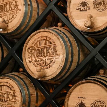 Stacked wooden whiskey barrels with branded logos for a cocktail and whiskey bar, displayed on a shelving unit.