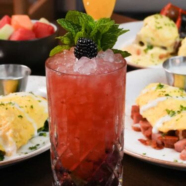 A vibrant red cocktail garnished with a blackberry and mint leaves, served in an old-fashioned glass with crushed ice. In the background, a plate of eggs benedict and a fruit bowl are