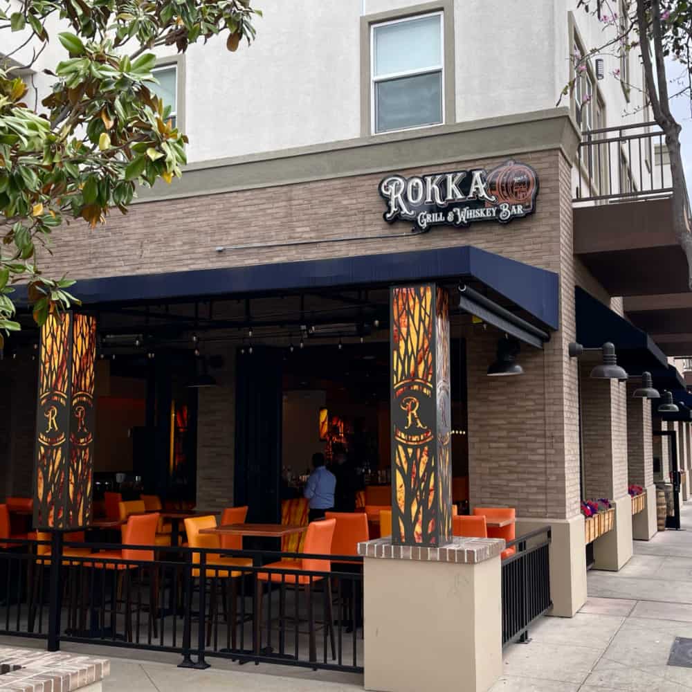 Exterior view of Rokka Grill & Whiskey Bar, featuring a covered dining area with orange chairs and decorative panels, situated on a street corner with trees. This whiskey bar is also famed for