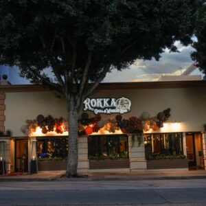 Facade of the rokkaku grill & parkway bar, illuminated by warm lighting at dusk and old fashioned jazz music playing in the background, is decorated with colorful balloons with a large tree at the front