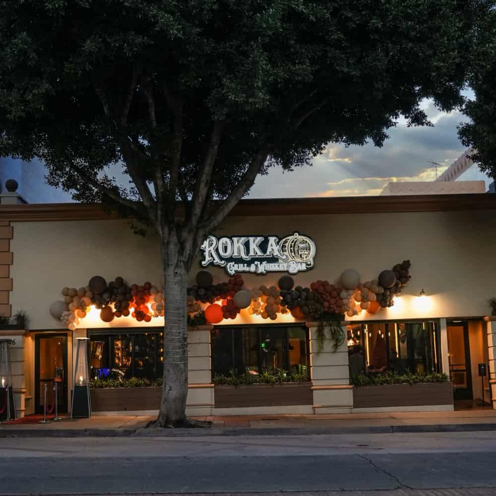 Facade of the rokkaku grill & parkway bar, illuminated by warm lighting at dusk and old fashioned jazz music playing in the background, is decorated with colorful balloons with a large tree at the front