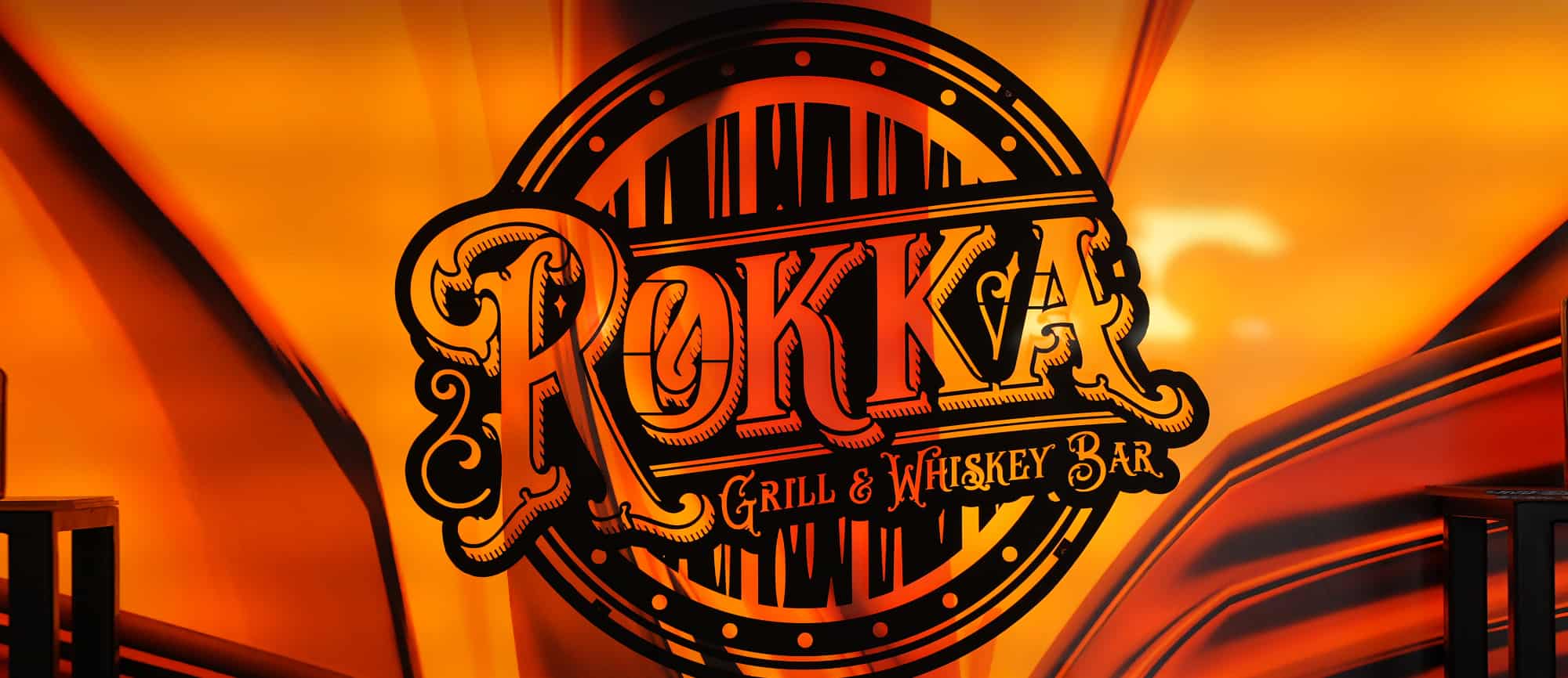 A vibrant sign for "rockar grill & whiskey bar" with elaborate black and orange graphics, set against a fiery orange backdrop that radiates outward, invites whiskey experts to enjoy a sophisticated experience.